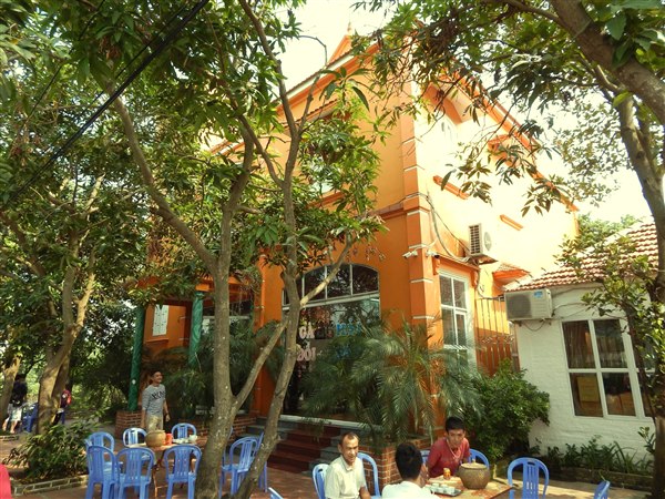 Sân trước nhà hàng Thăng Long ngày trước: nhà sơn cam dưới tán cây, khách ngồi uống trà ở bộ bàn ghế nhựa